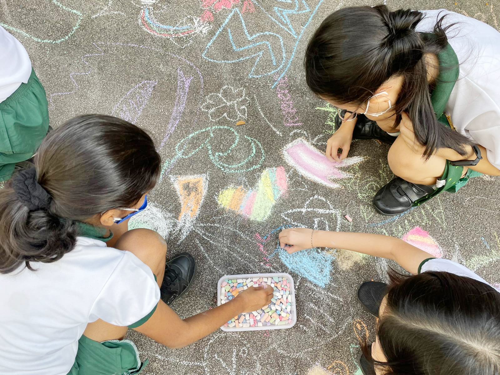 Students kneel on the ground creating colourful chalk drawings together on the pavement during an outdoor art activity.